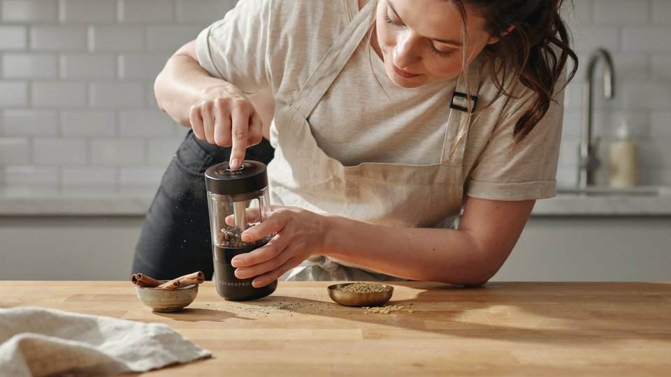 Woman demonstrating how to use a spice grinder on a kitchen counter with whole spices like peppercorns and cinnamon nearby.