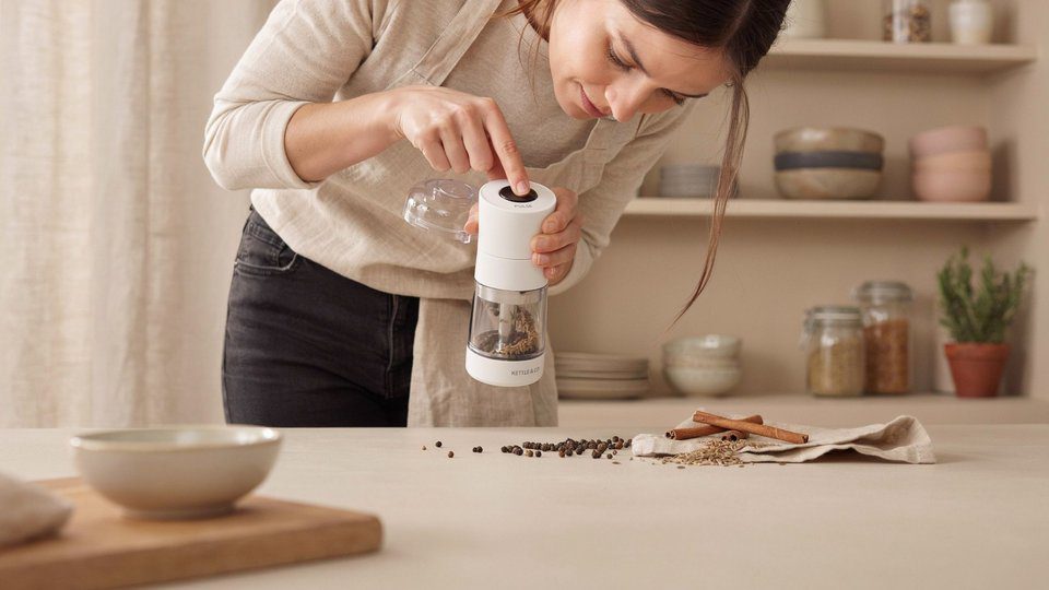 Woman in a kitchen carefully operating a spice grinder with whole spices placed on the counter for grinding.