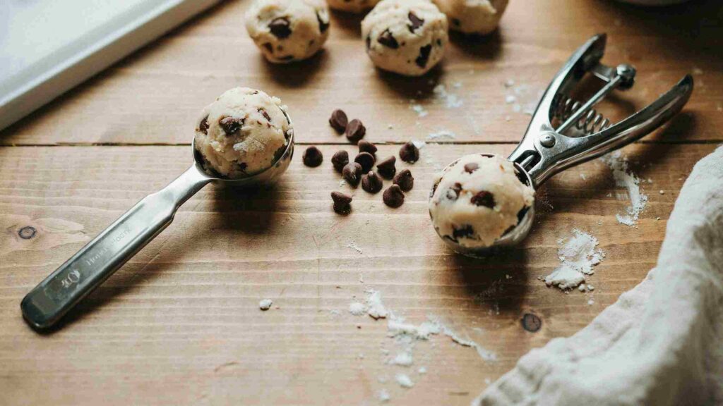 A baking sheet of cookies showing uneven sizes and browning from tablespoon portioning next to a row of uniform, evenly baked cookies from a cookie scoop, comparing cookie scoop vs tablespoon results.