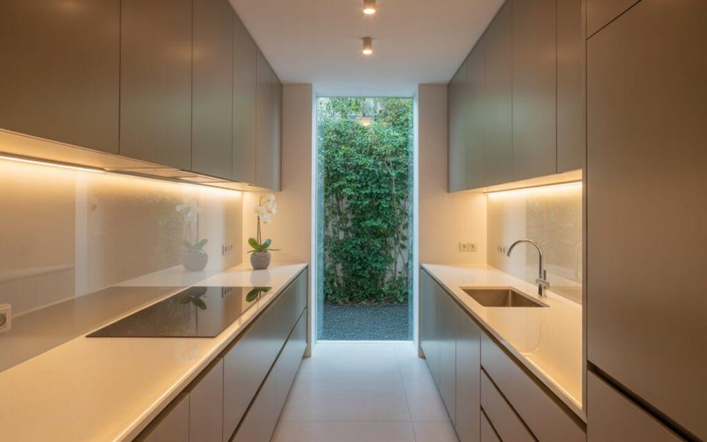 A sleek modern galley kitchen with under-cabinet lighting and a floor-to-ceiling glass window offering a lush garden view